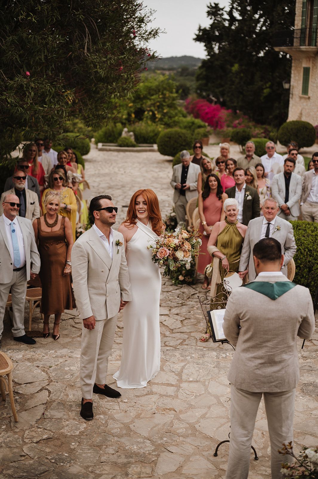 Bride and groom stand together before officiant during sunny outdoor wedding ceremony with guests watching.