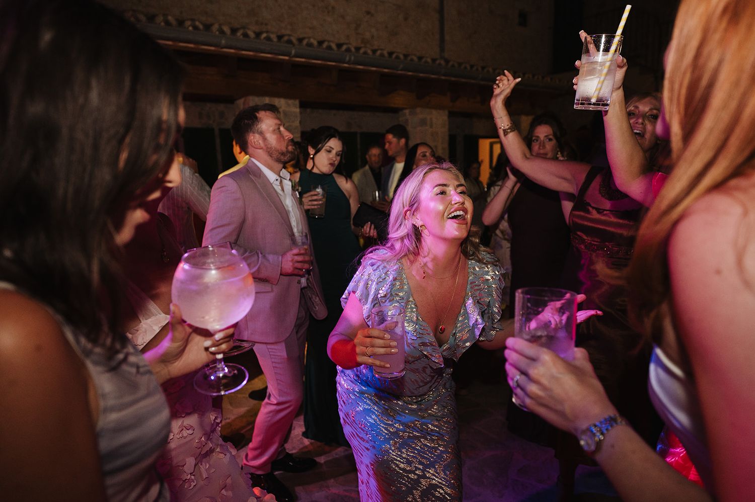 Woman in silver sequin dress laughing and dancing under pink lights at wedding party.