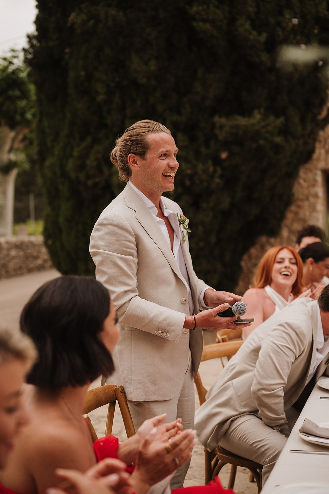 Smiling groom in cream suit holding microphone while giving speech at outdoor wedding.