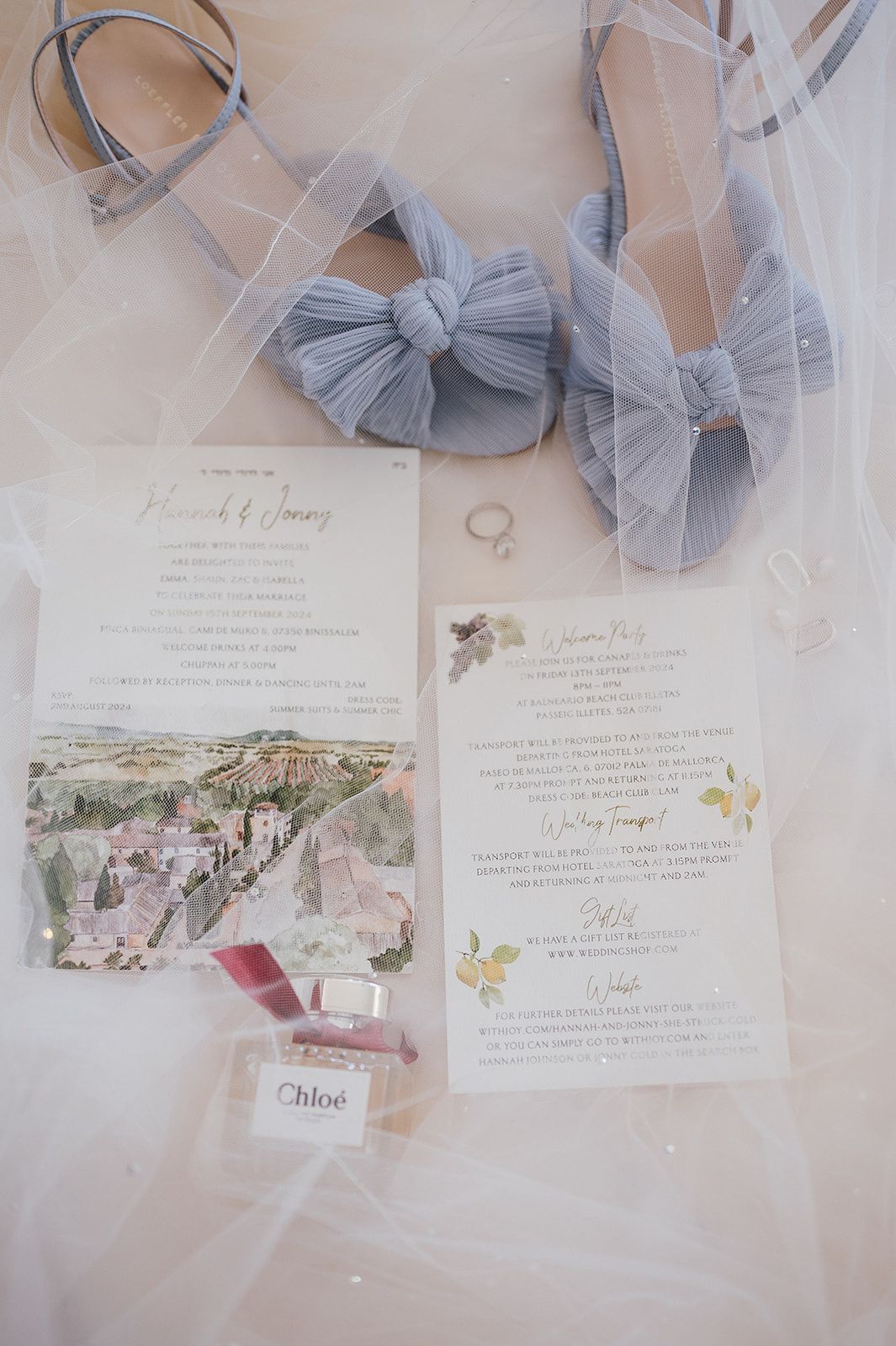 Wedding flat lay featuring blue bow heels, floral invitations, engagement ring, and Chloe perfume on veil.