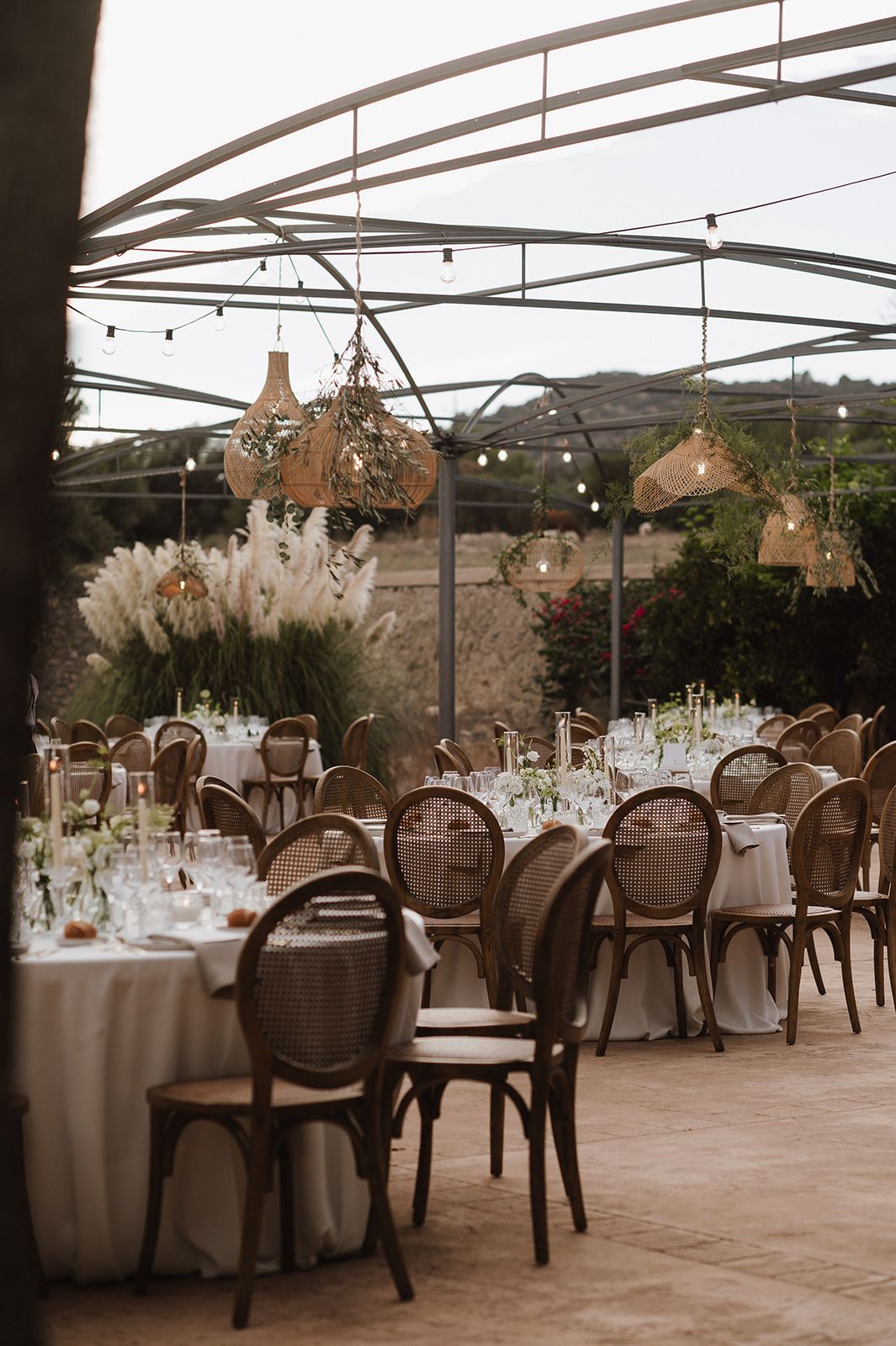 Elegant outdoor wedding reception under glass canopy with rattan pendant lights and round tables.