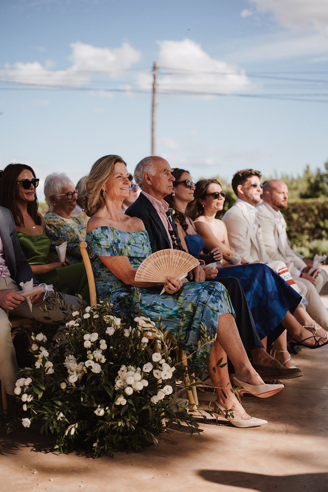 Wedding guests seated outdoors in sunlight, with a woman in blue floral dress holding a decorative fan.