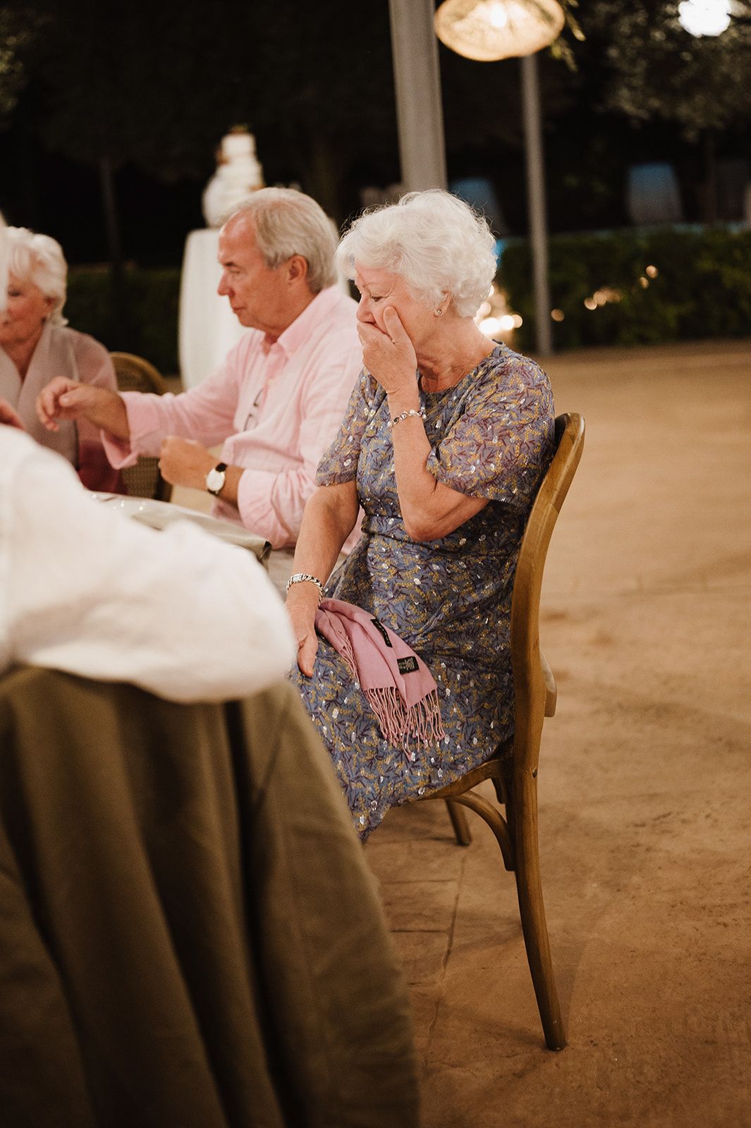 Elderly woman covering her mouth with hand while seated at outdoor evening wedding reception table.
