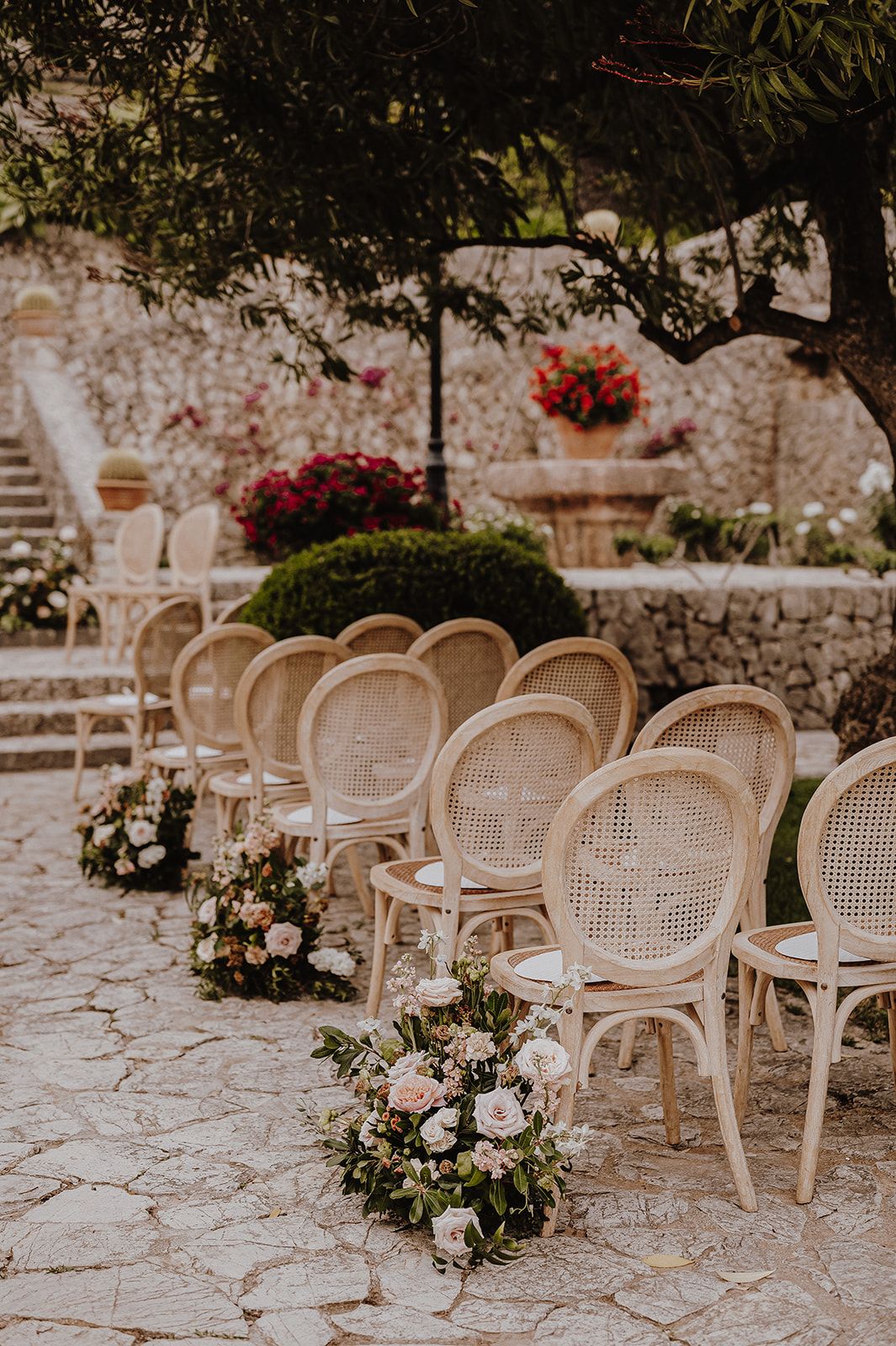 Elegant rattan wedding ceremony chairs lined with blush floral arrangements in rustic garden venue.