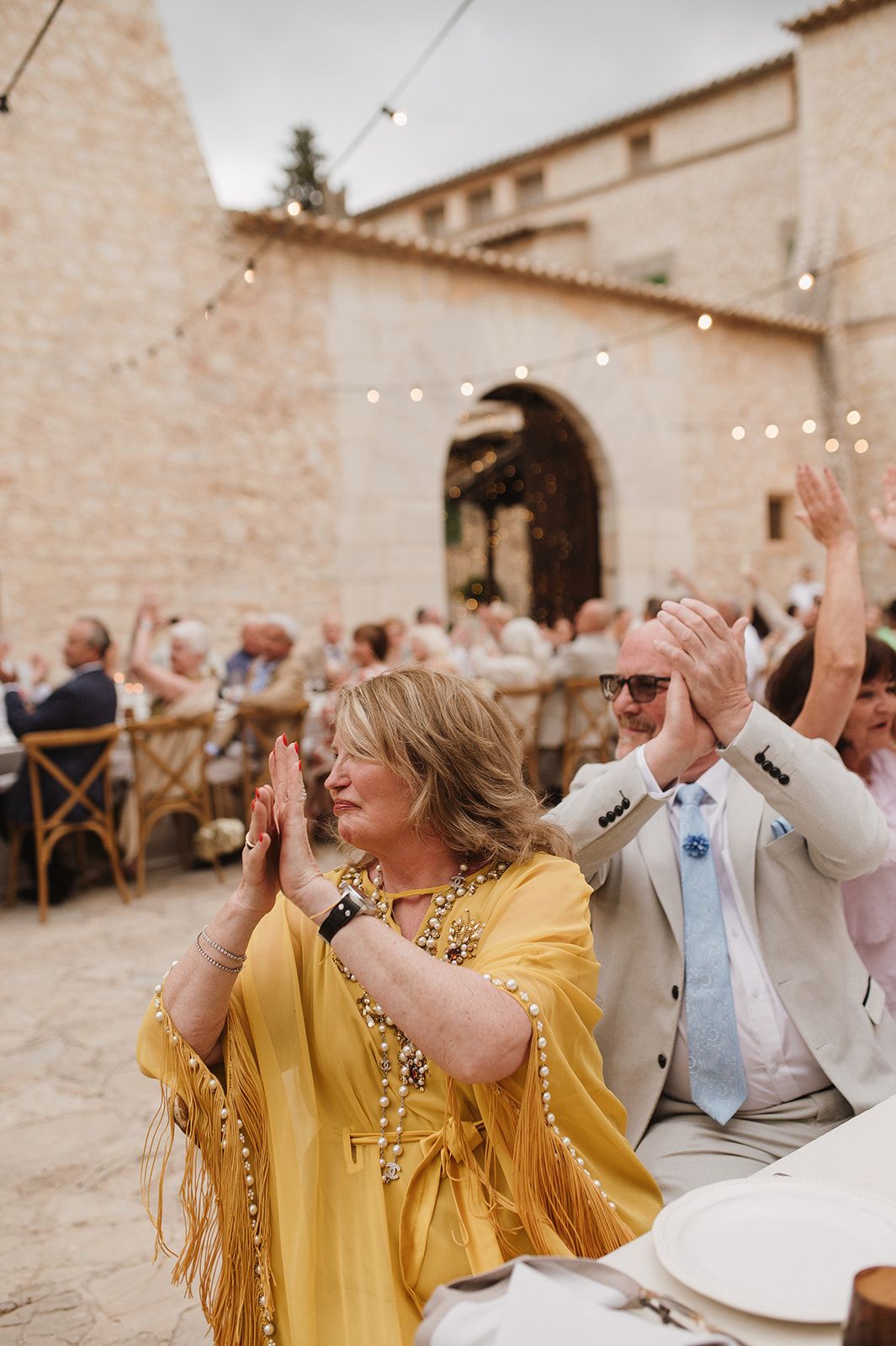 Wedding guests in yellow fringed dress and beige suit clap enthusiastically at an outdoor stone venue reception.