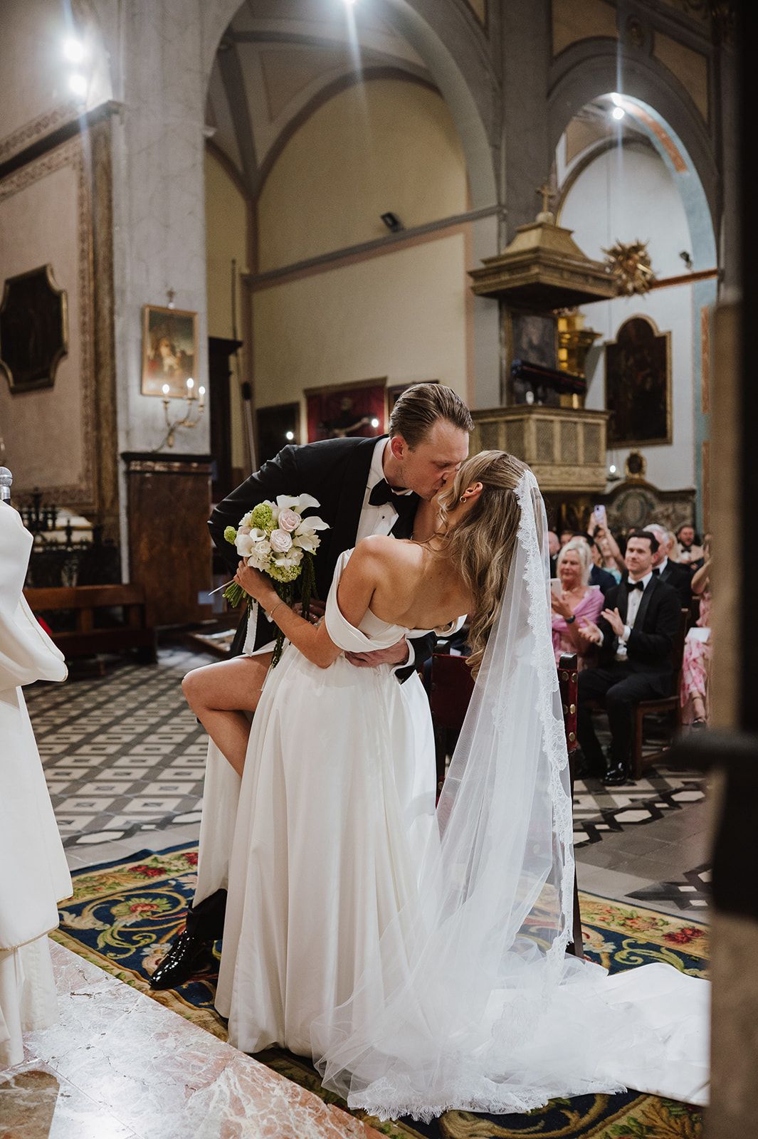 Bride and groom share first kiss in ornate church aisle as guests applaud during wedding ceremony.