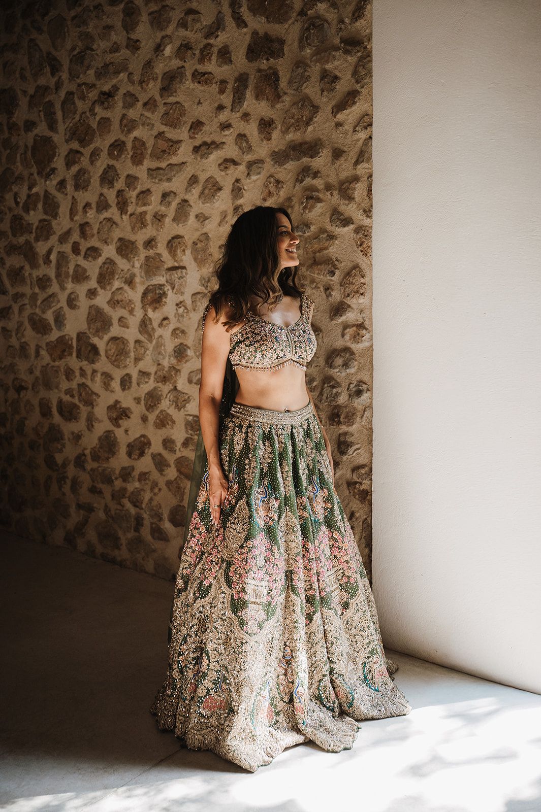 Bride in ornate green floral lehenga posing against rustic stone wall.
