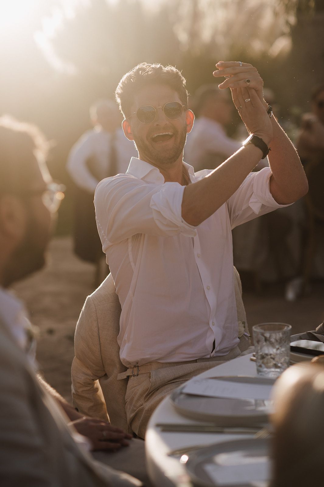 Smiling man in white shirt raises his hand at a sunlit outdoor wedding reception table at golden hour.
