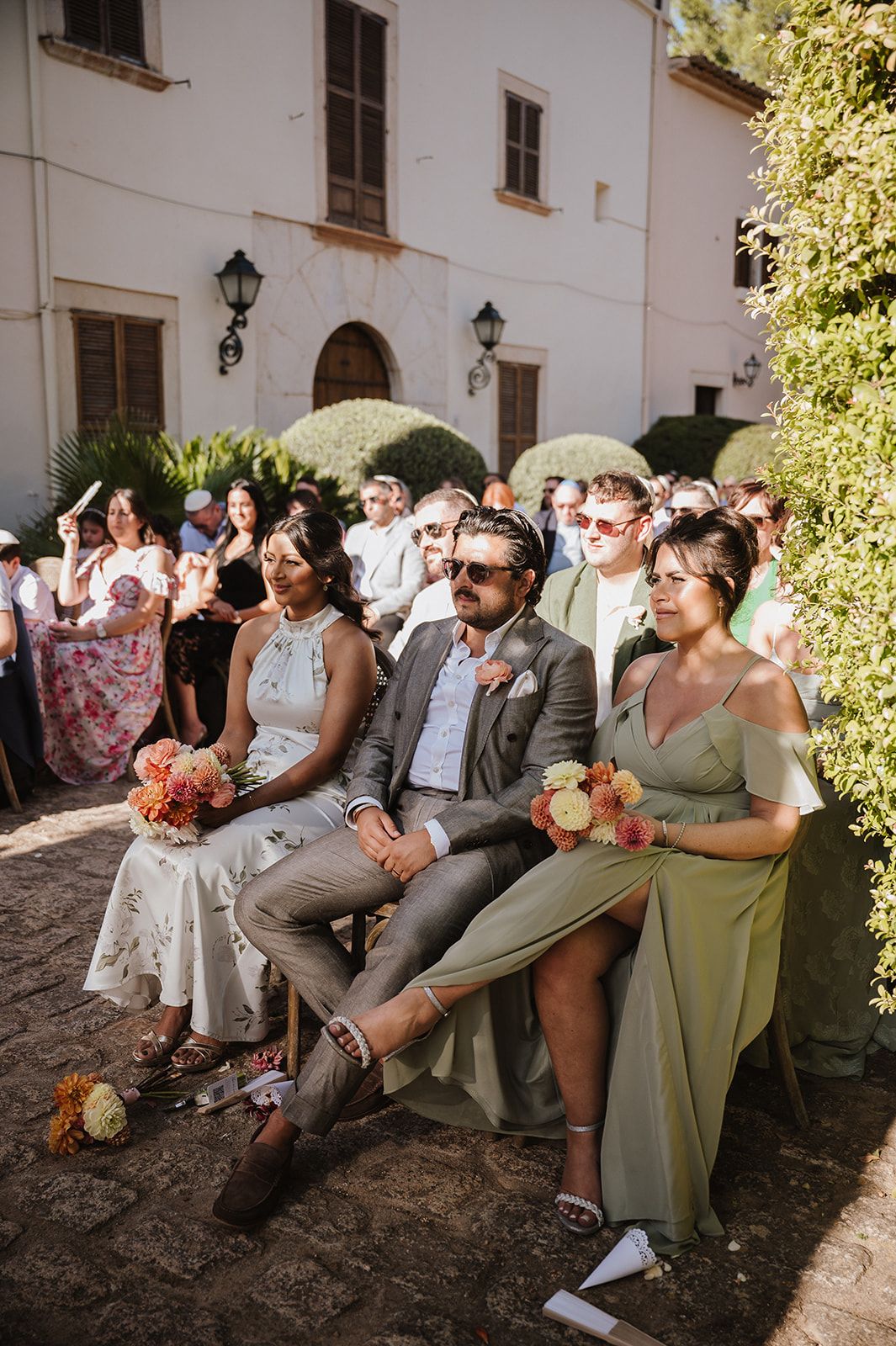Wedding guests seated outdoors in sunglasses watching a sunny ceremony at a rustic European venue.