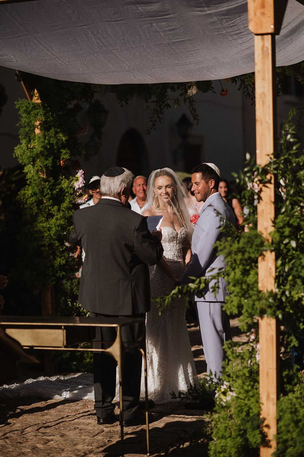 Bride and groom exchange vows under a floral chuppah during an outdoor Jewish wedding ceremony.