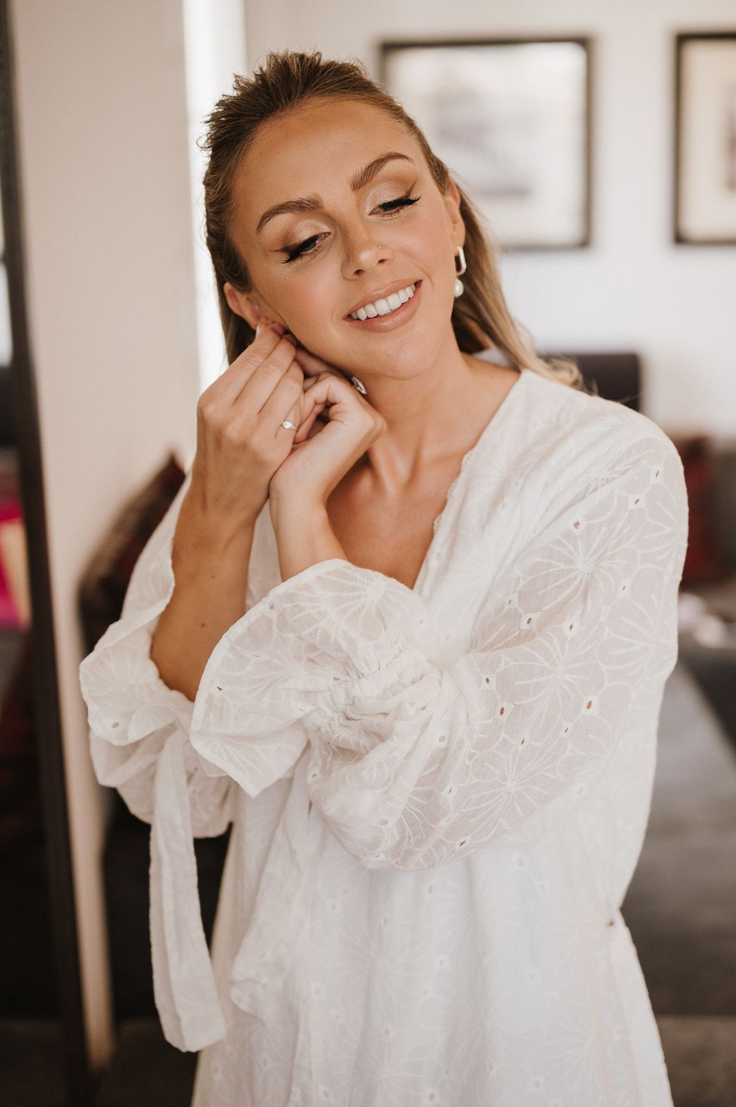 Bride in white lace robe smiling while putting on earrings during wedding preparations.
