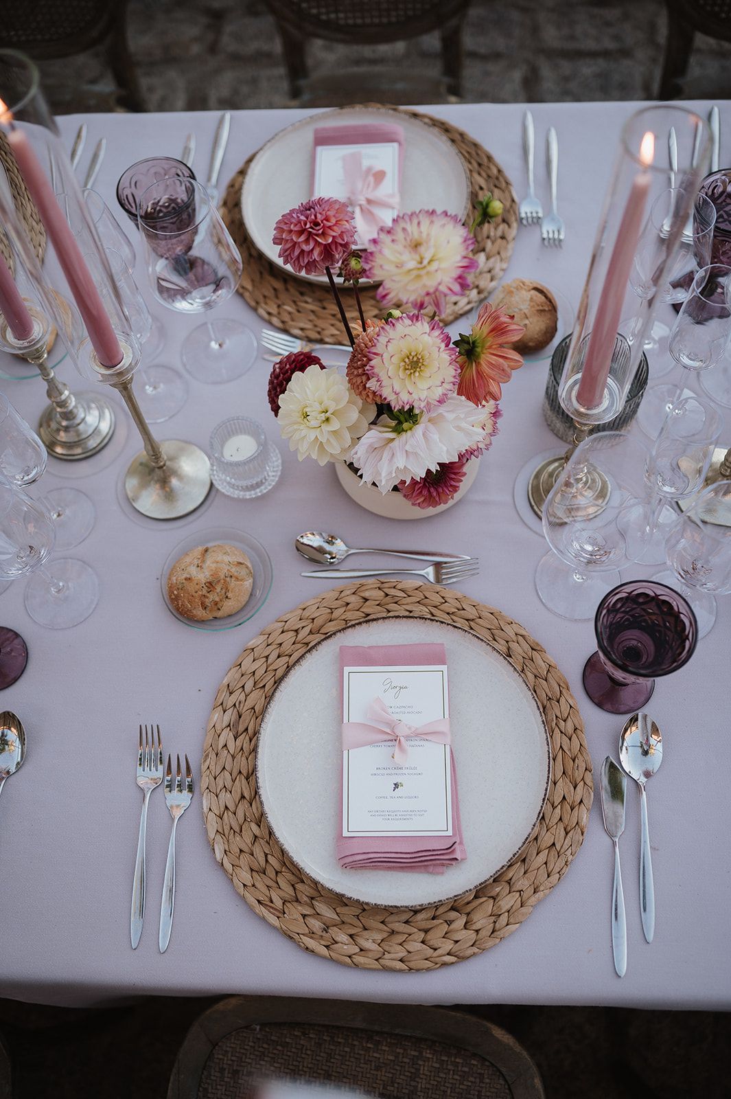 Elegant wedding table setting with pink dahlias, tapered candles, woven chargers, and purple glasses.