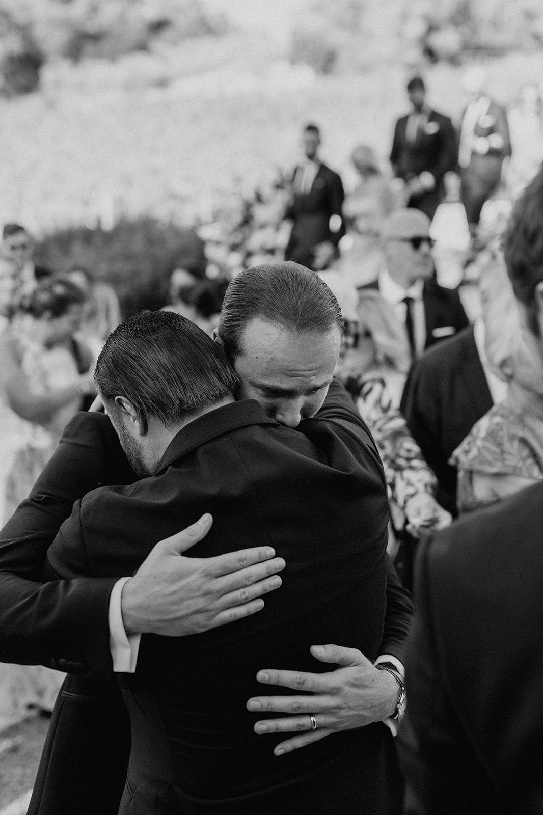 Two men in suits share an emotional hug at an outdoor wedding ceremony.