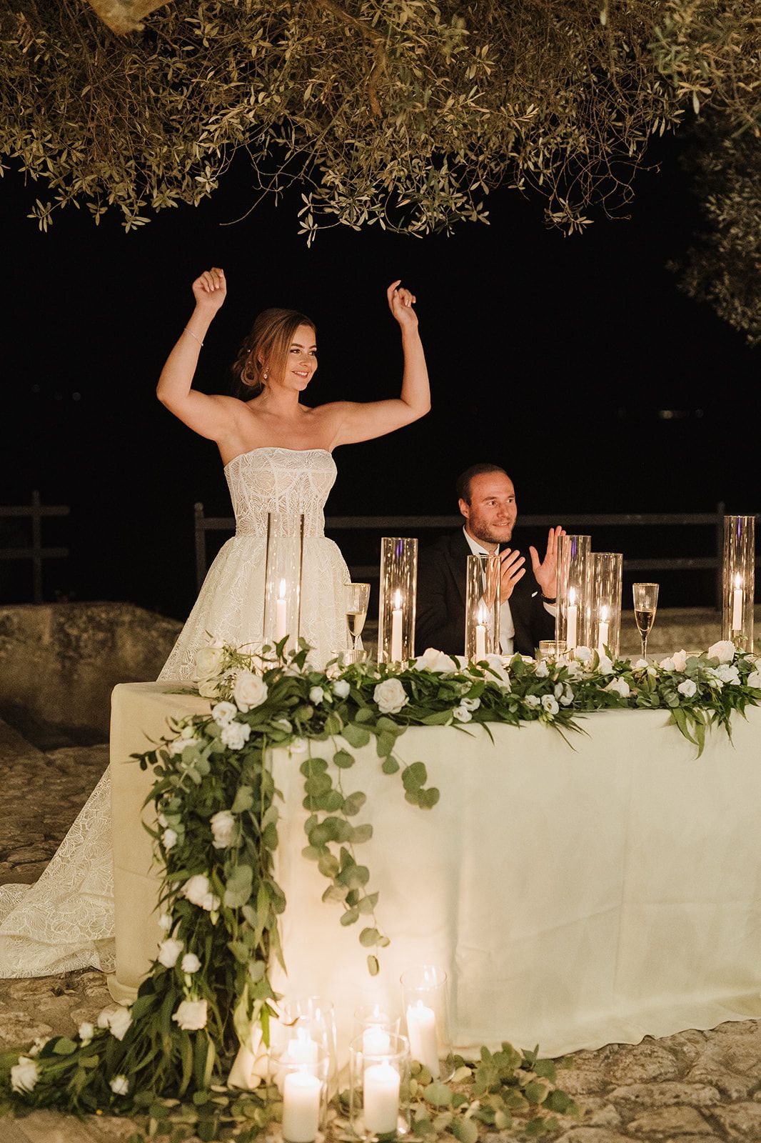 Bride raising arms in celebration at candlelit sweetheart table beneath olive tree at night.