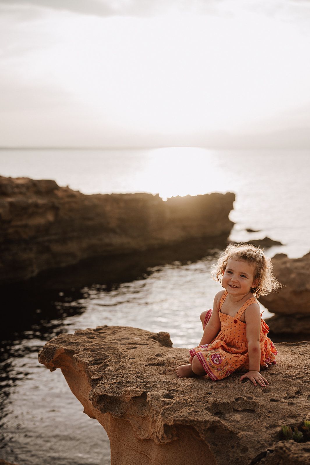 Smiling toddler girl in orange dress sitting on coastal rocks at golden sunset.