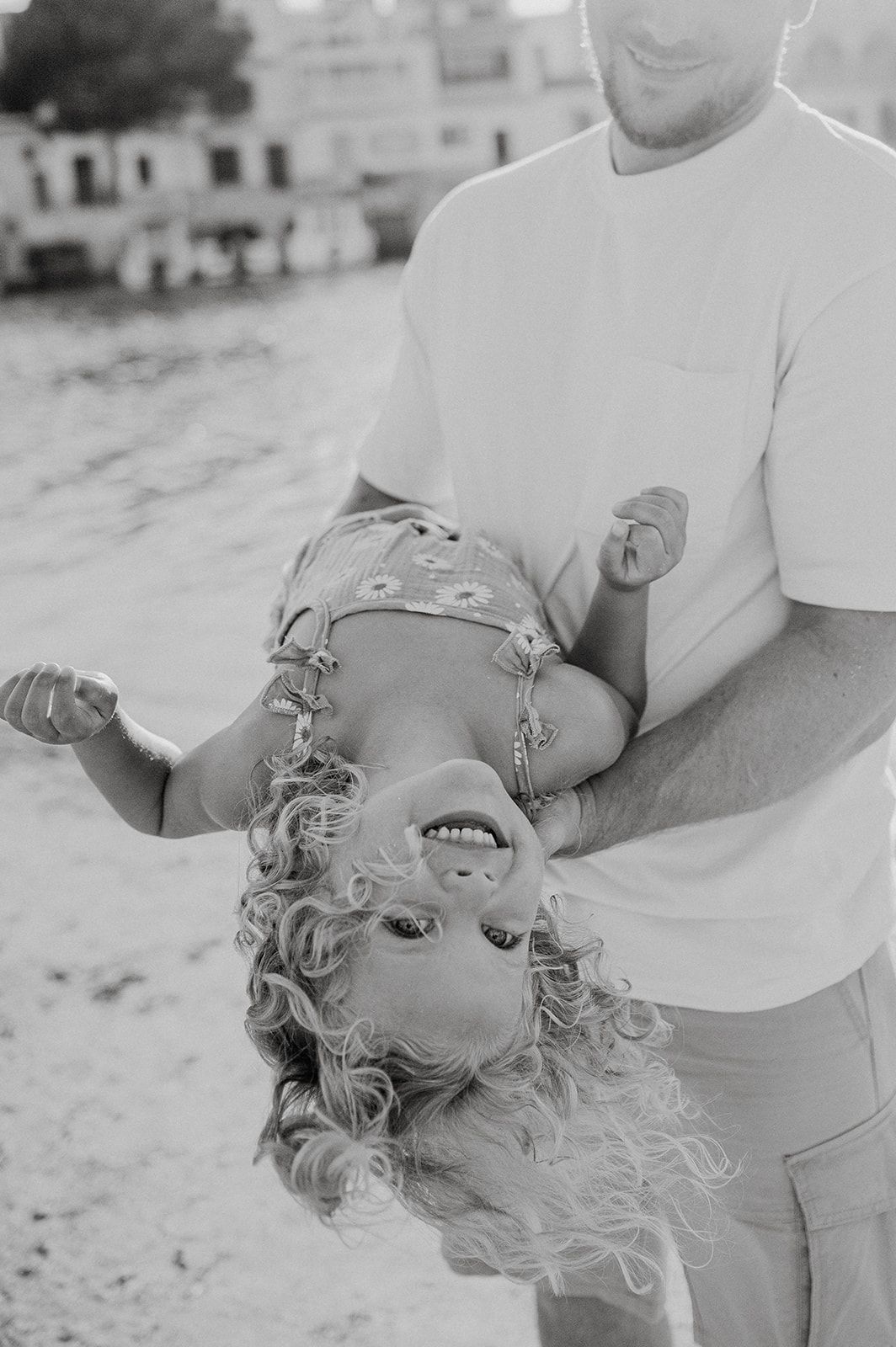 Father playfully holds his curly-haired child upside down on a sunny beach in black and white photo.