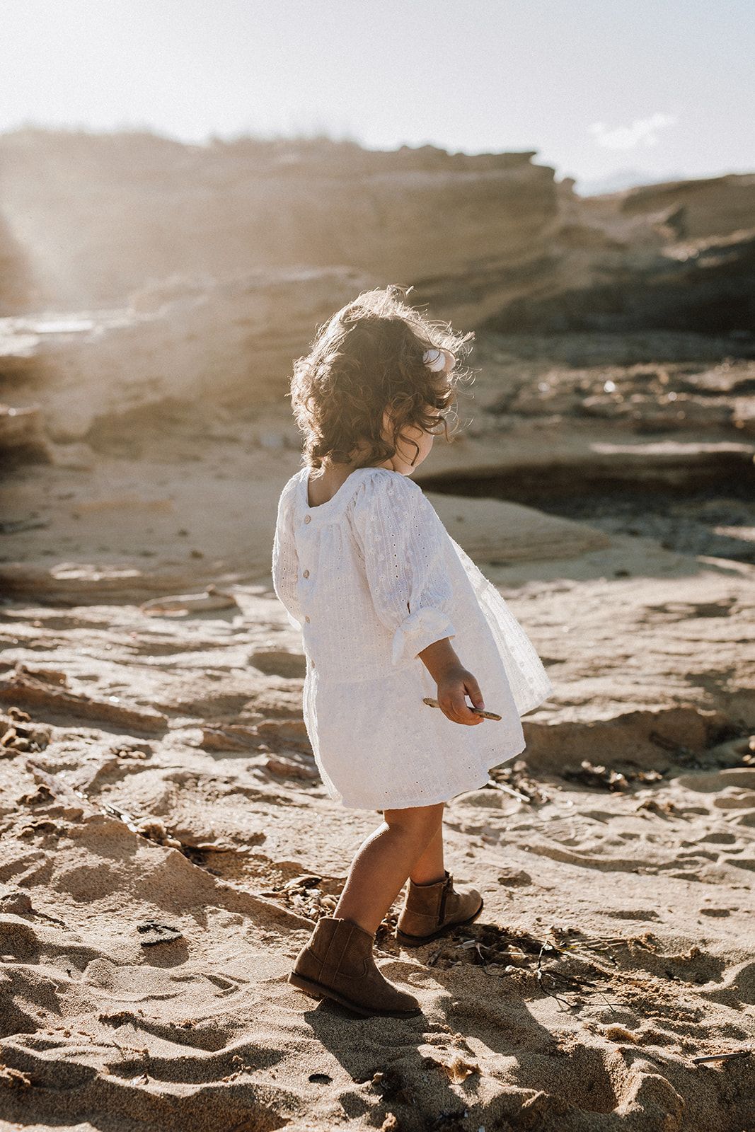A curly-haired toddler girl in a white dress and brown boots walks on a sandy sunlit beach.