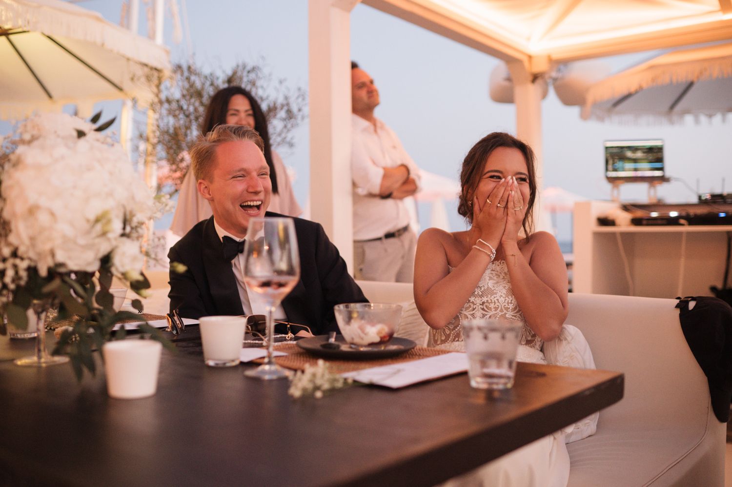 Bride and groom laugh joyfully during wedding reception speeches at an elegant oceanfront venue.