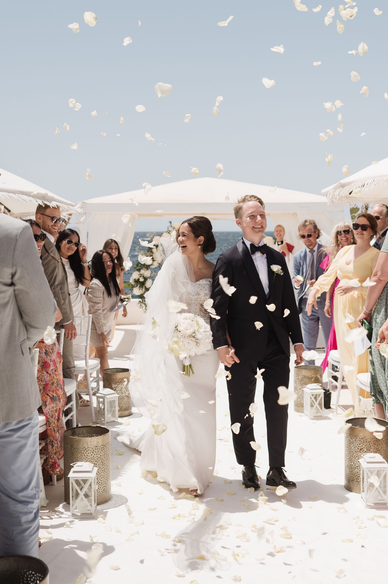 Bride and groom walk down aisle as white rose petals shower them at seaside wedding.