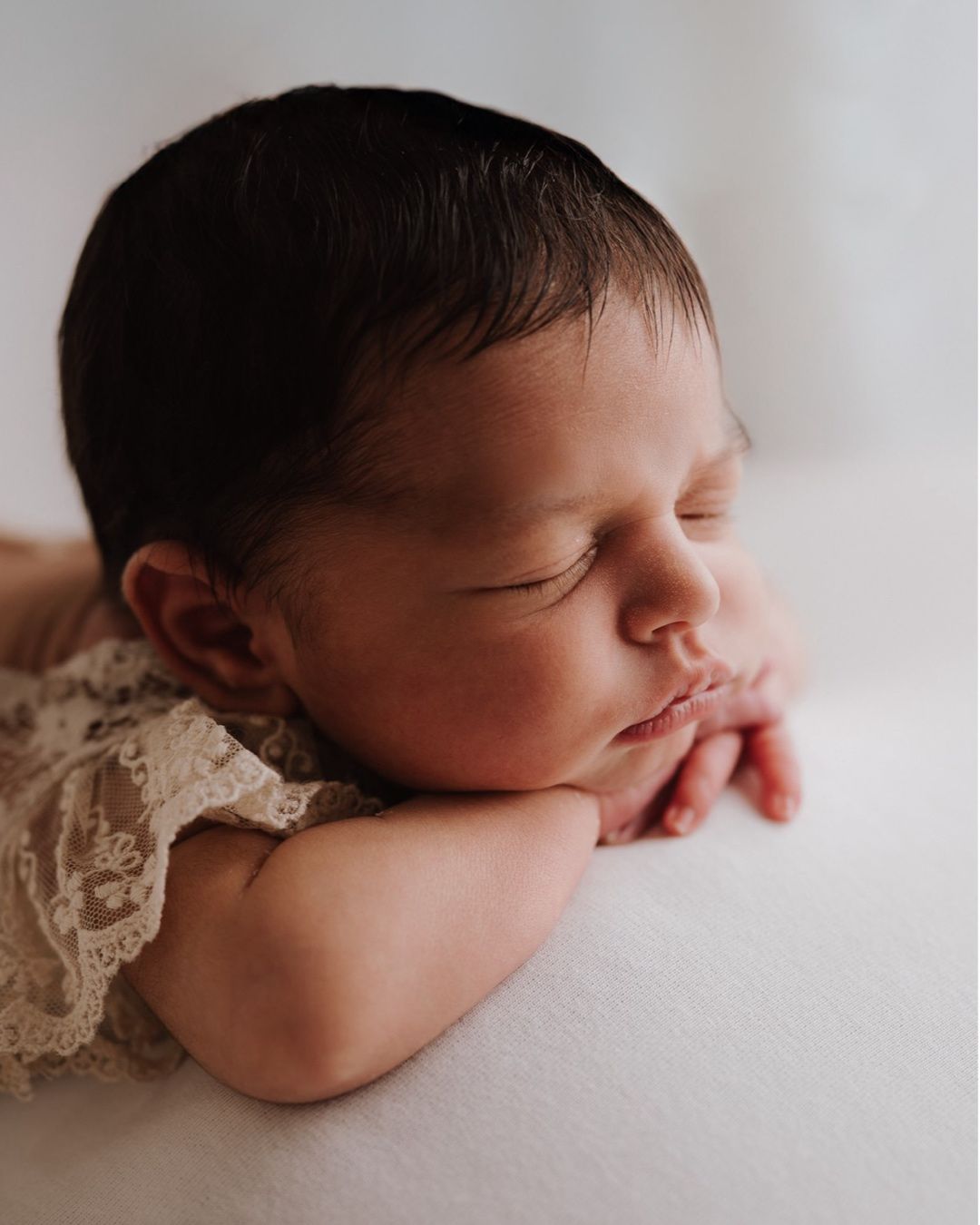 Peaceful newborn baby sleeping with hands under cheek, wearing lace romper, soft white background.