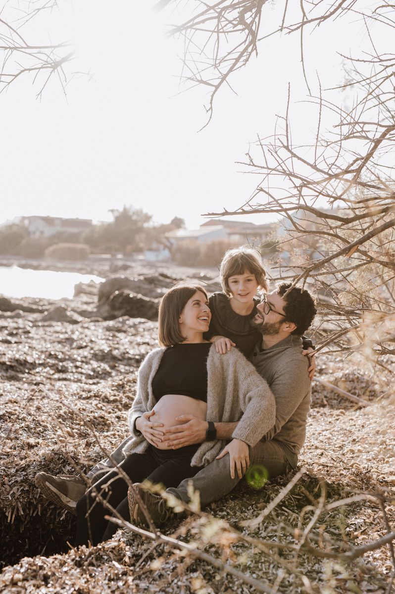 Pregnant woman sits outdoors with partner and child in warm sunlight near rocky shore.