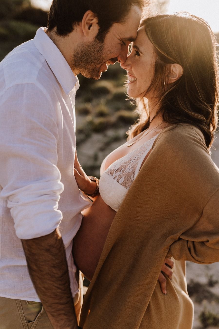 A couple shares a tender moment at golden hour, with the woman showing a baby bump in a warm outdoor maternity photo session.