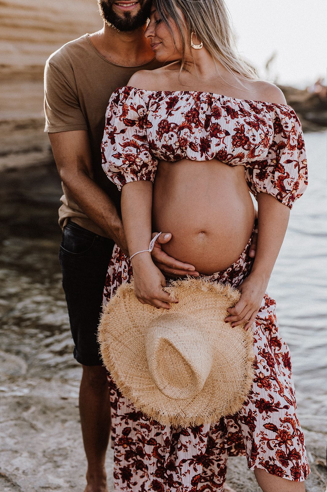 Close-up of pregnant couple embracing on beach, woman in floral crop top showcasing baby bump.