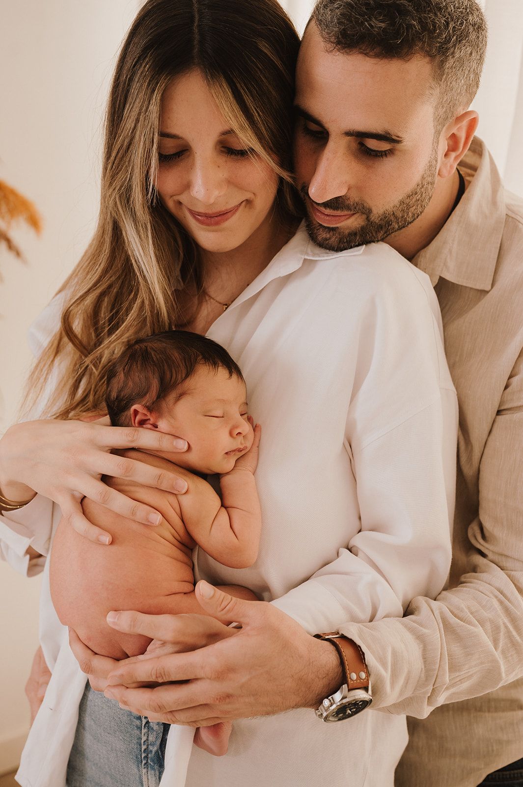 A couple tenderly holds their sleeping newborn baby, smiling warmly in a soft, natural light portrait.