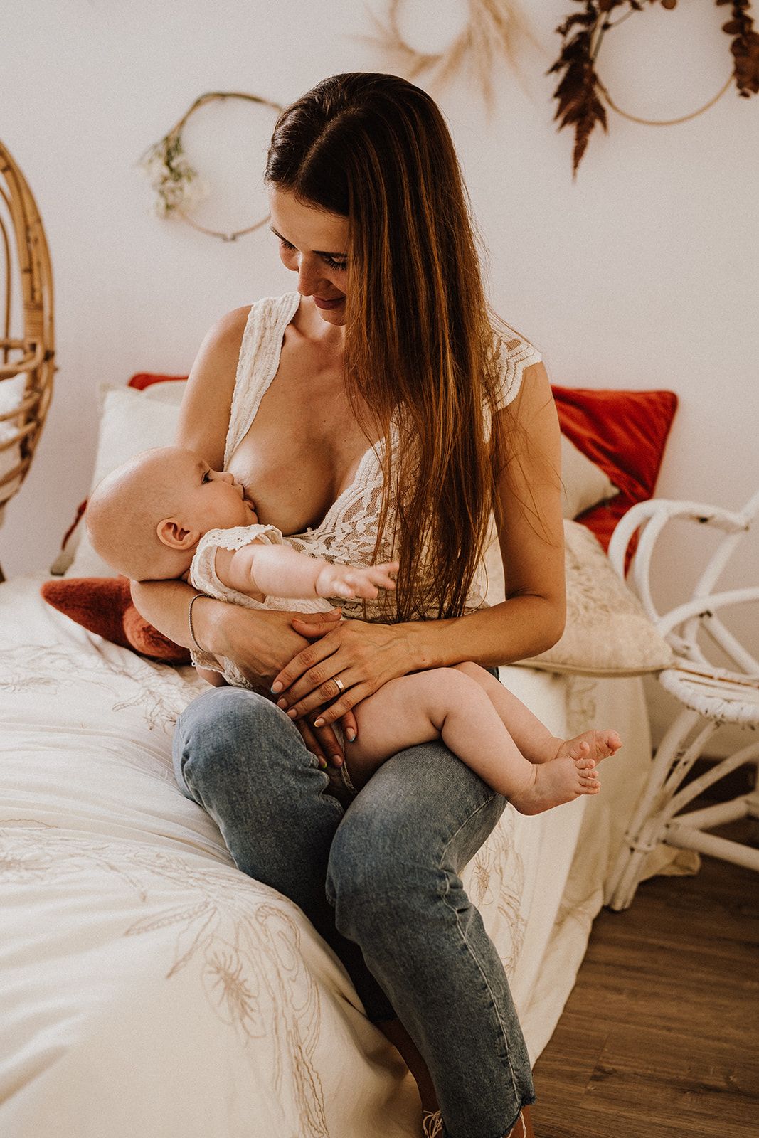 A smiling mother breastfeeding her newborn baby while sitting on a bed in a cozy boho-style bedroom.