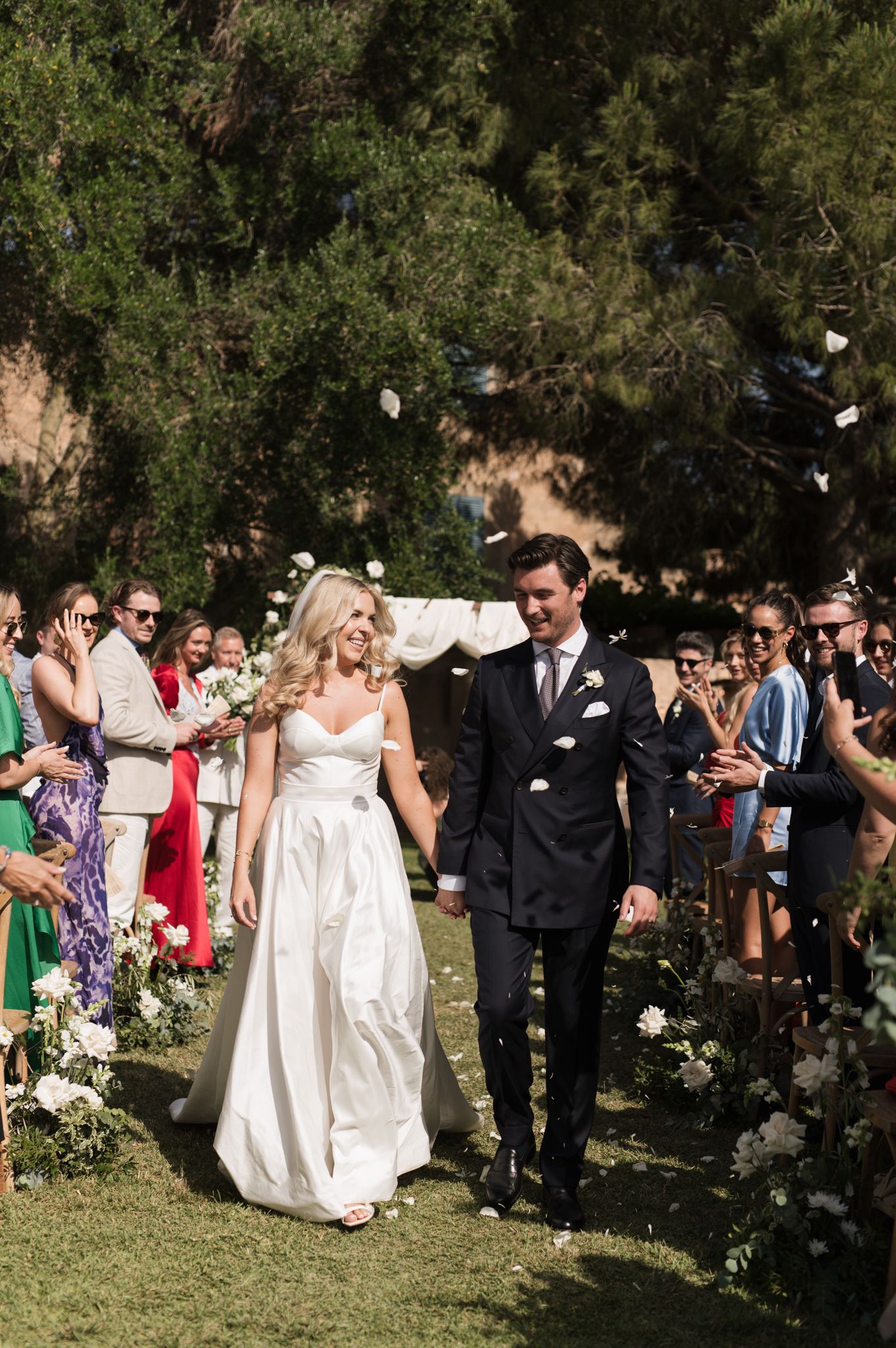 Newlyweds walk down the aisle as guests toss petals in celebration at an outdoor garden wedding ceremony.