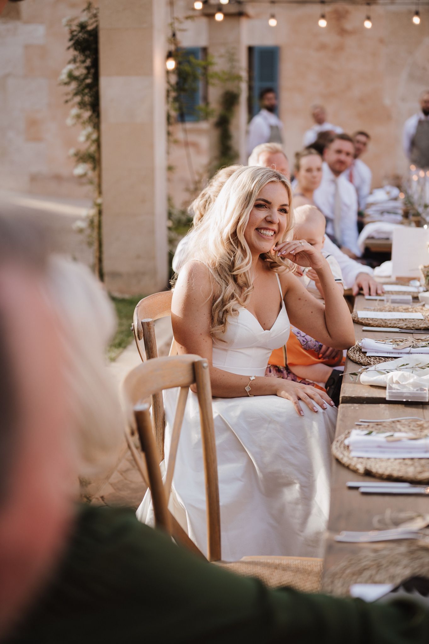 Radiant blonde bride in white gown smiling at guests during an outdoor wedding reception dinner.