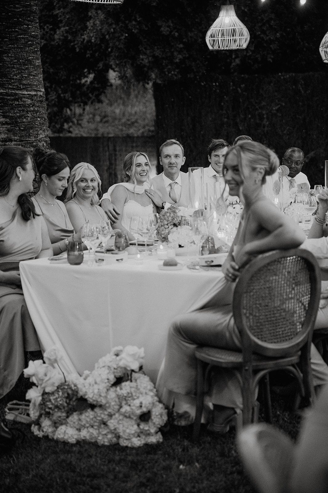 Black and white photo of wedding party laughing at candlelit outdoor reception dinner table.