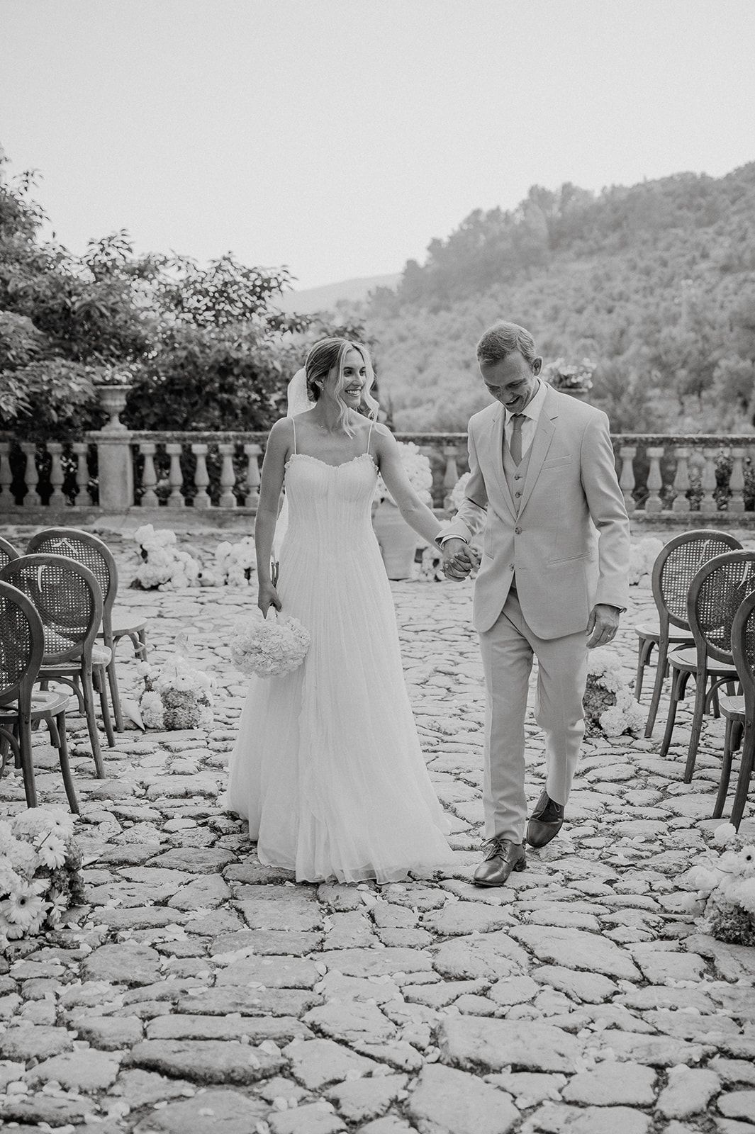 Bride and groom walk hand in hand down cobblestone aisle after outdoor wedding ceremony in black and white.