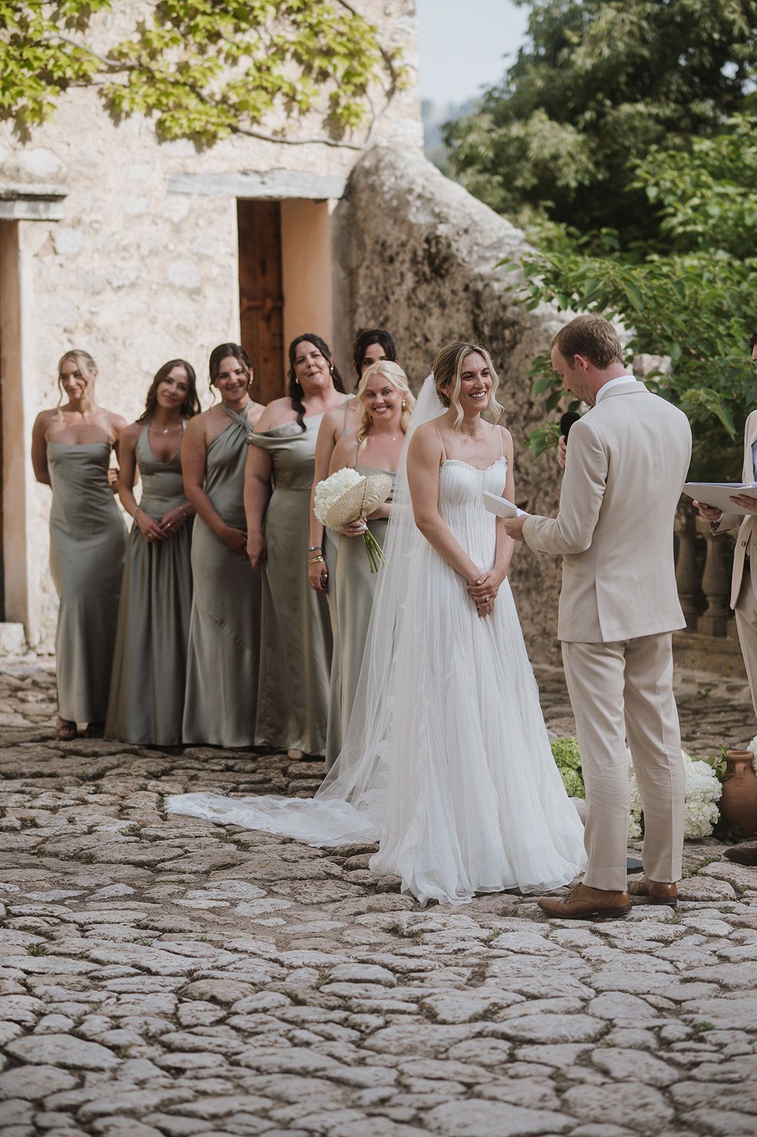 Bride with white bouquet stands with groom as bridesmaids in sage green dresses line up beside them.