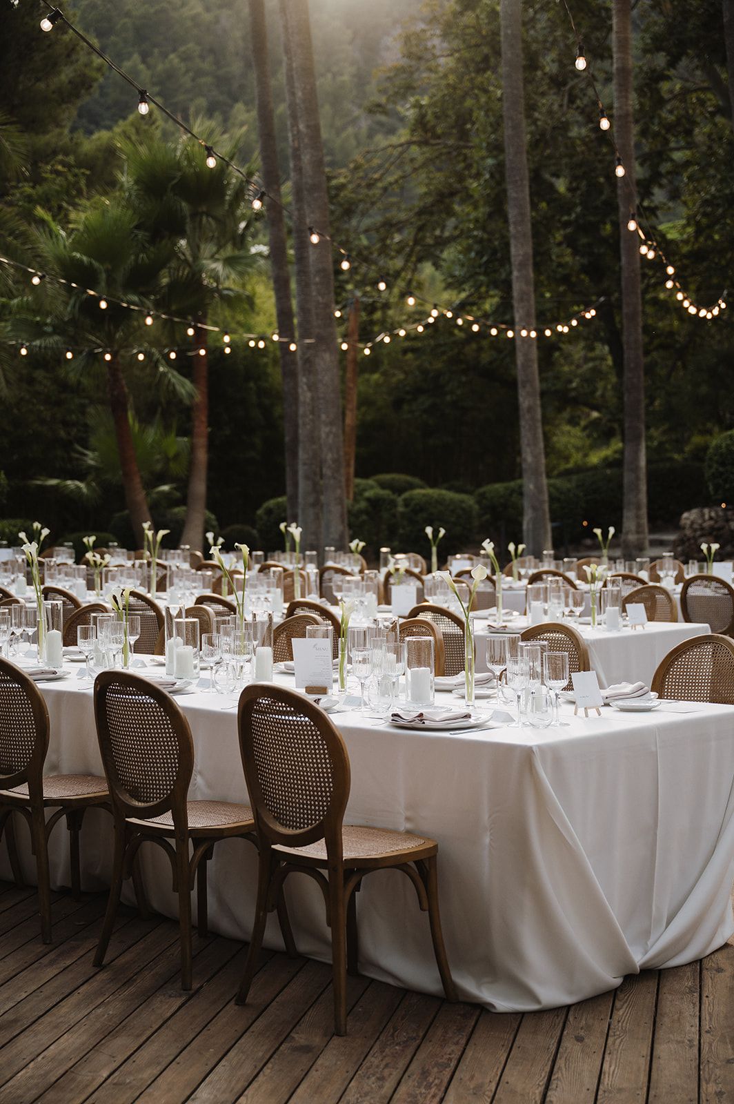 Long outdoor wedding reception tables with white linens, cane chairs, and warm string lights at sunset.
