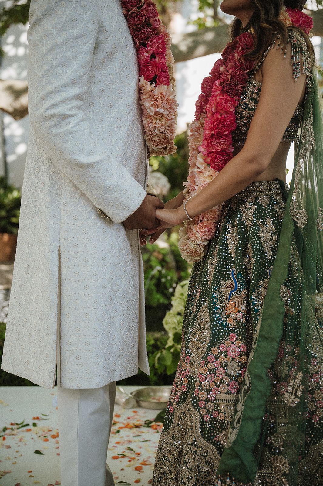 Bride in embroidered green lehenga and groom in white sherwani hold hands during an Indian wedding ceremony.