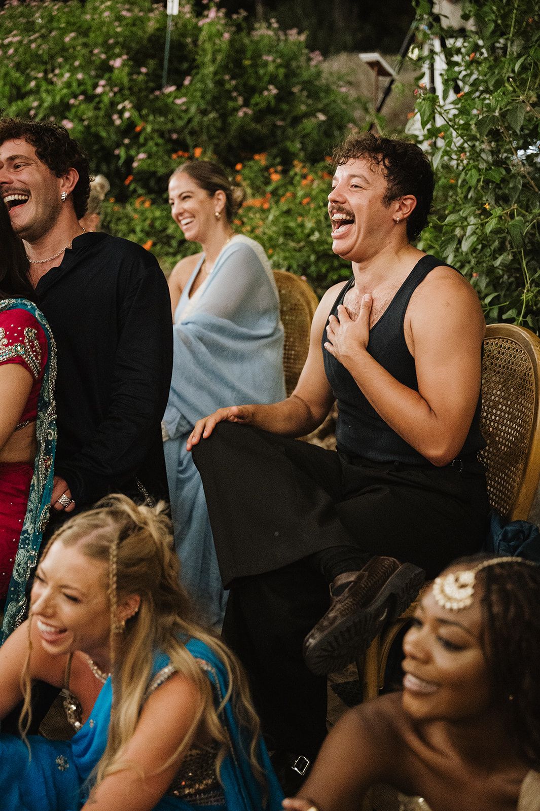 Guests laughing joyfully during Indian wedding reception speeches in lush outdoor garden setting.