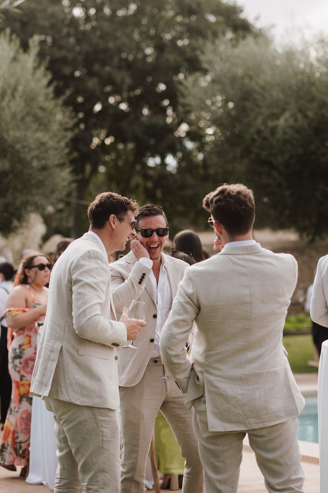 Three men in linen suits laugh together poolside at an outdoor wedding reception.
