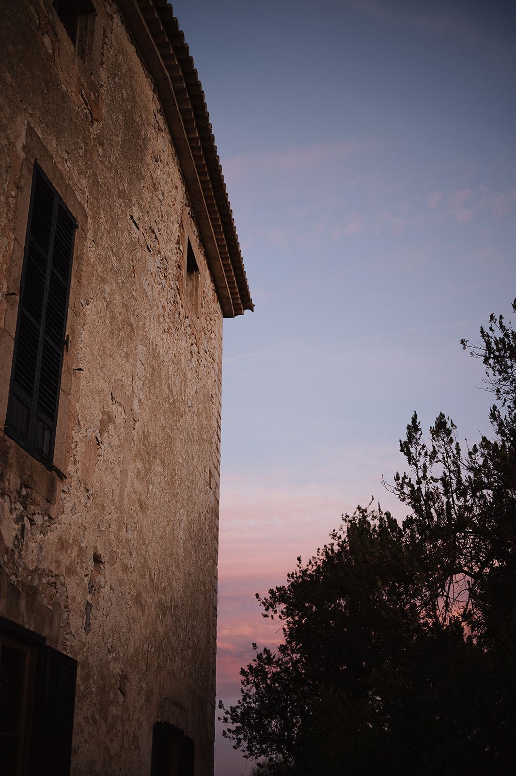 Old stone building with dark windows against a pink and blue twilight sky, with silhouetted trees in the background.