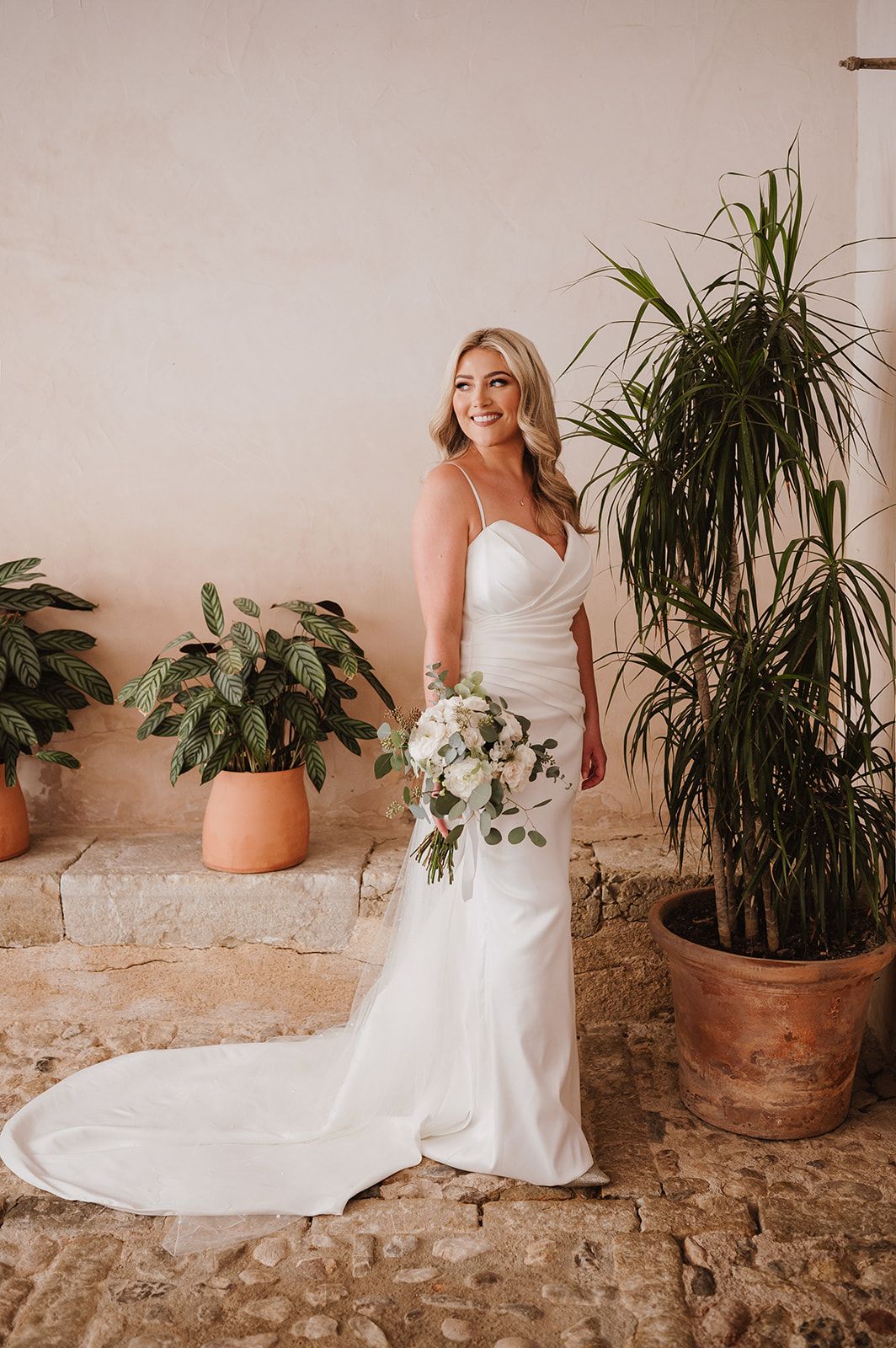Smiling bride in a strapless white fitted wedding gown holding a white bouquet surrounded by tropical potted plants.