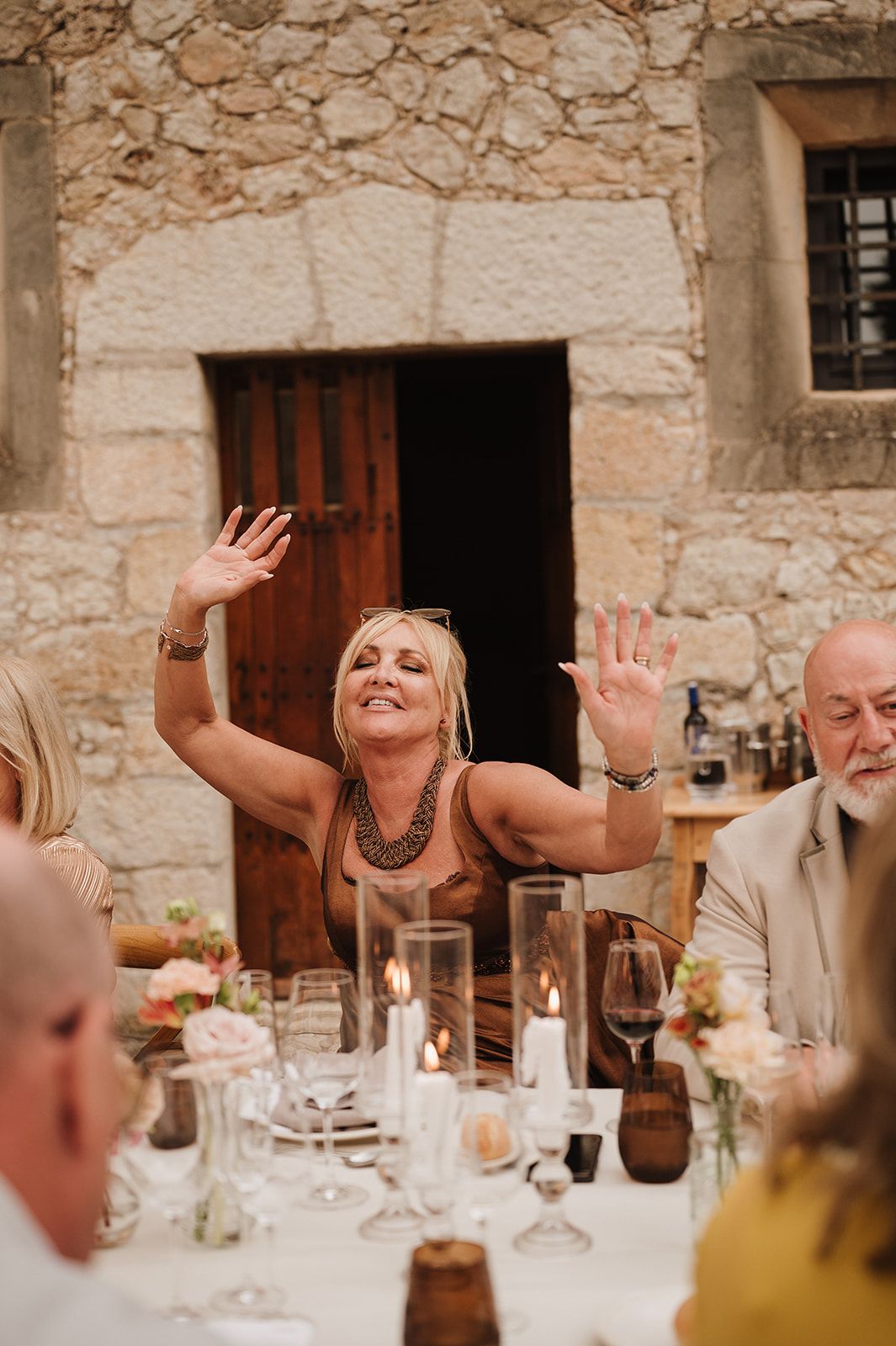 Joyful wedding guest raises hands expressively at candlelit outdoor stone courtyard reception dinner.