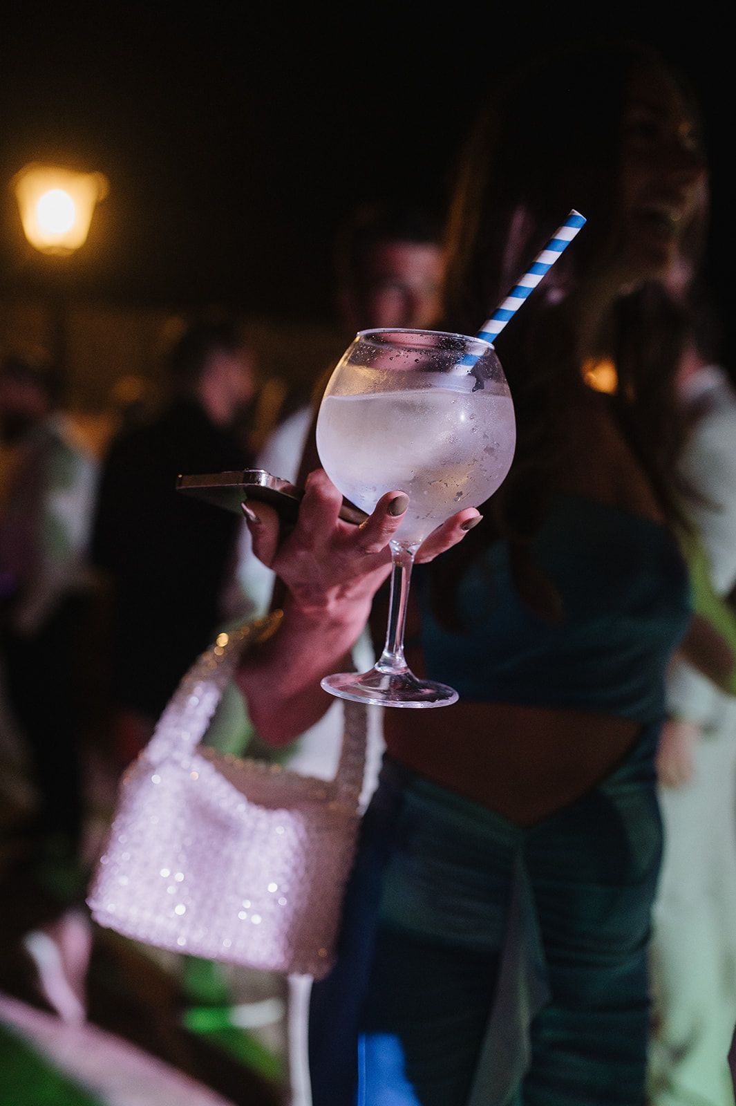 Hand holding a large gin and tonic cocktail glass with blue straw at an outdoor wedding party.