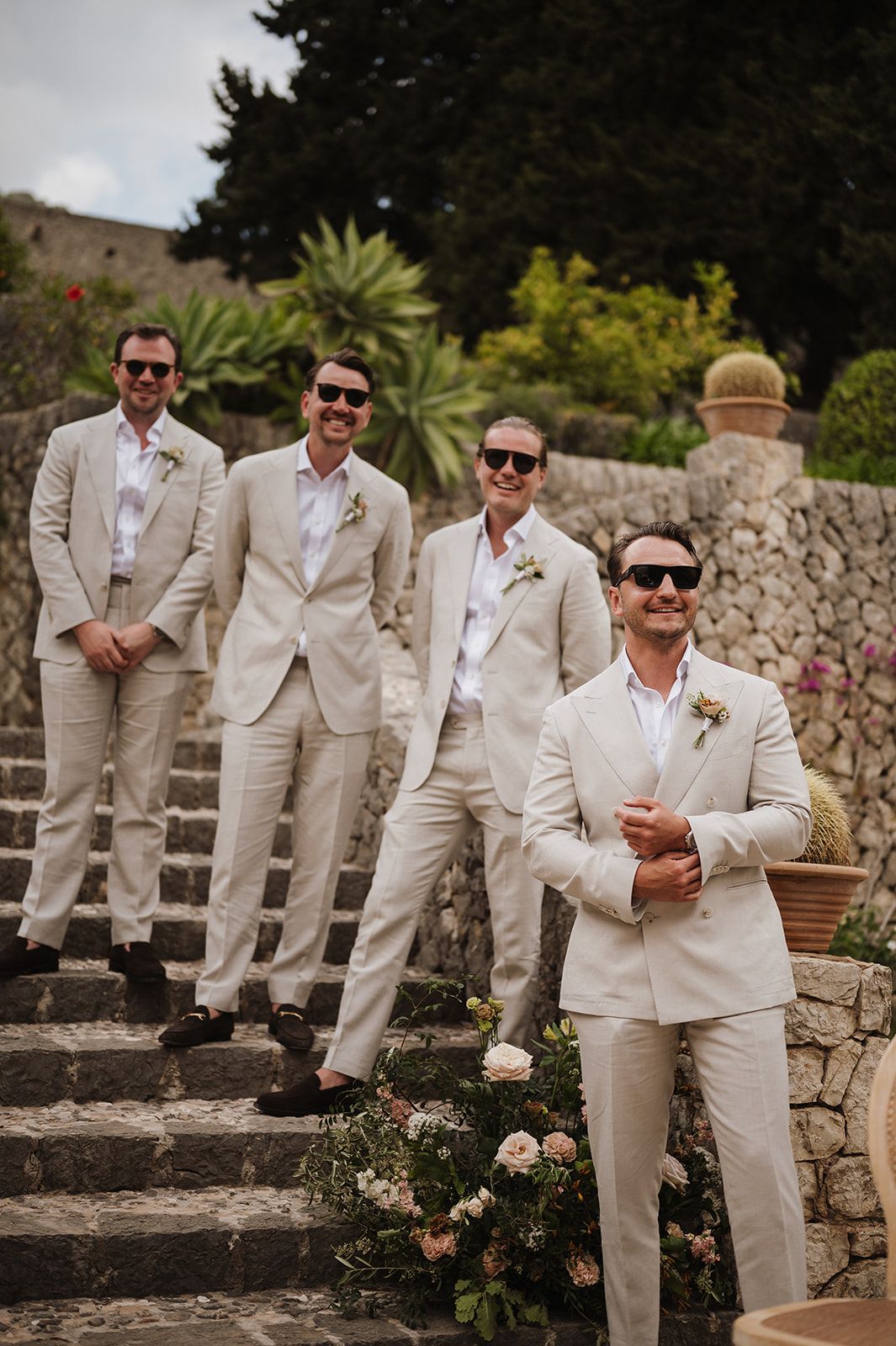 Four groomsmen in matching cream linen suits with boutonnieres pose on stone steps at wedding.