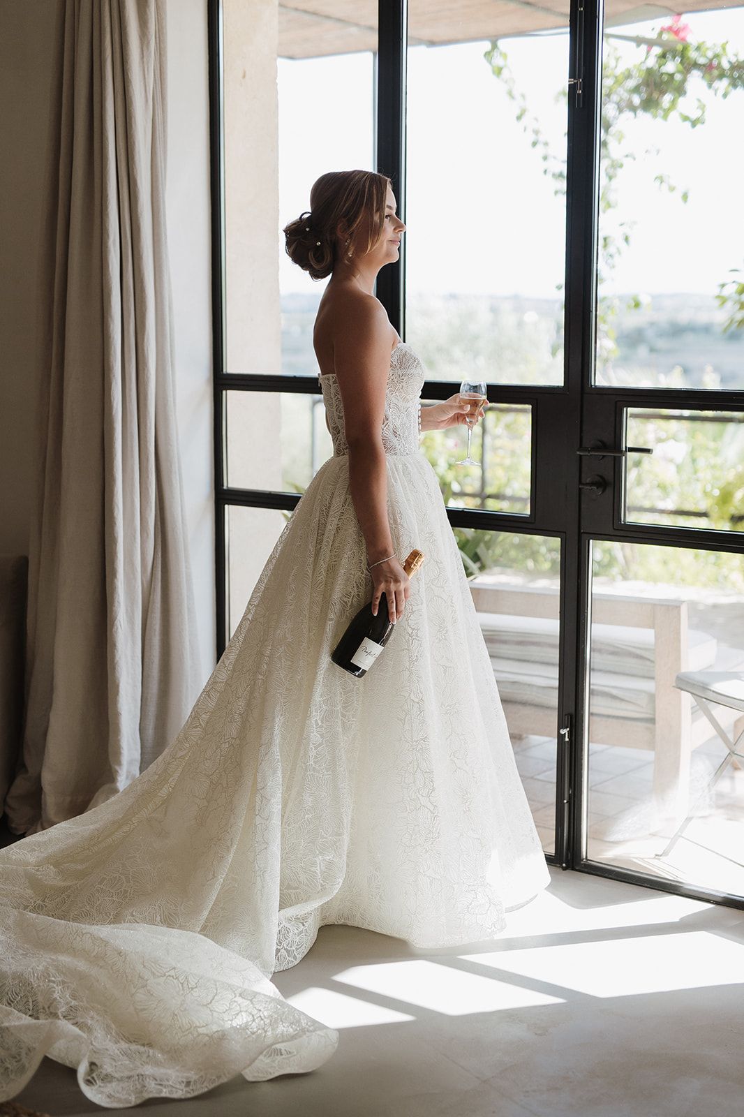 Bride in strapless lace ball gown holding champagne bottle and glass while gazing out a large window.