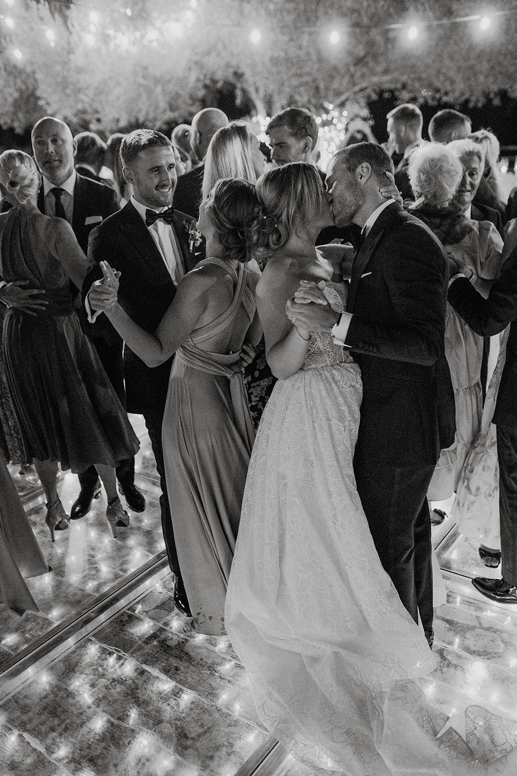 Black and white photo of bride and guests dancing together on lit dance floor at wedding reception.