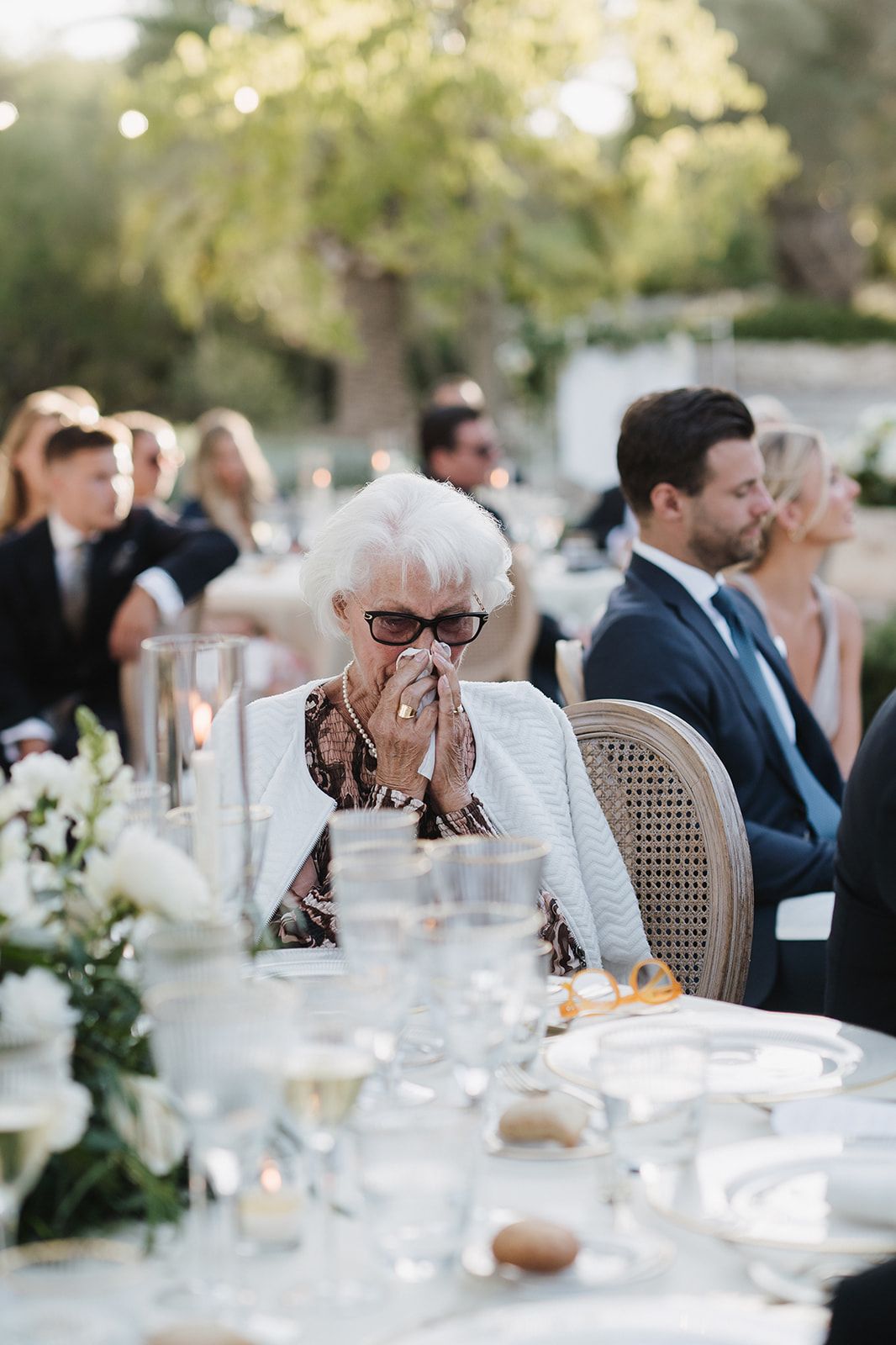 Elderly wedding guest with white hair and glasses wipes tears at an outdoor reception dinner table.