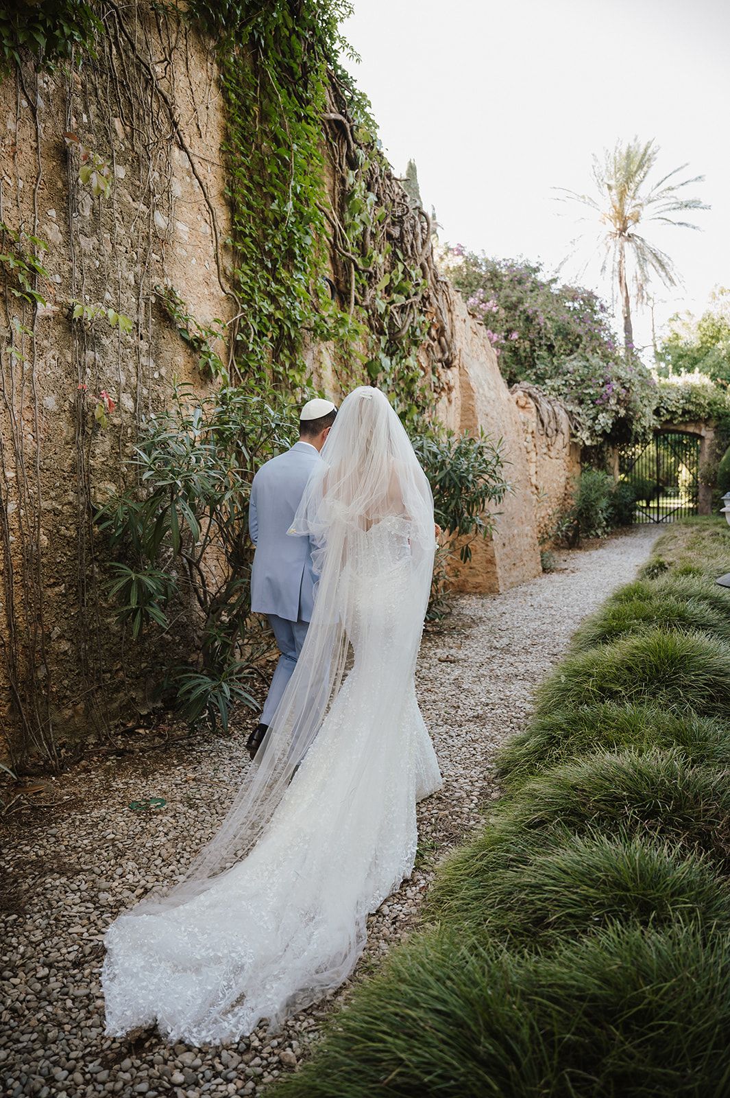 Bride in lace gown with long veil and groom in light suit walking along ivy-covered stone garden path.