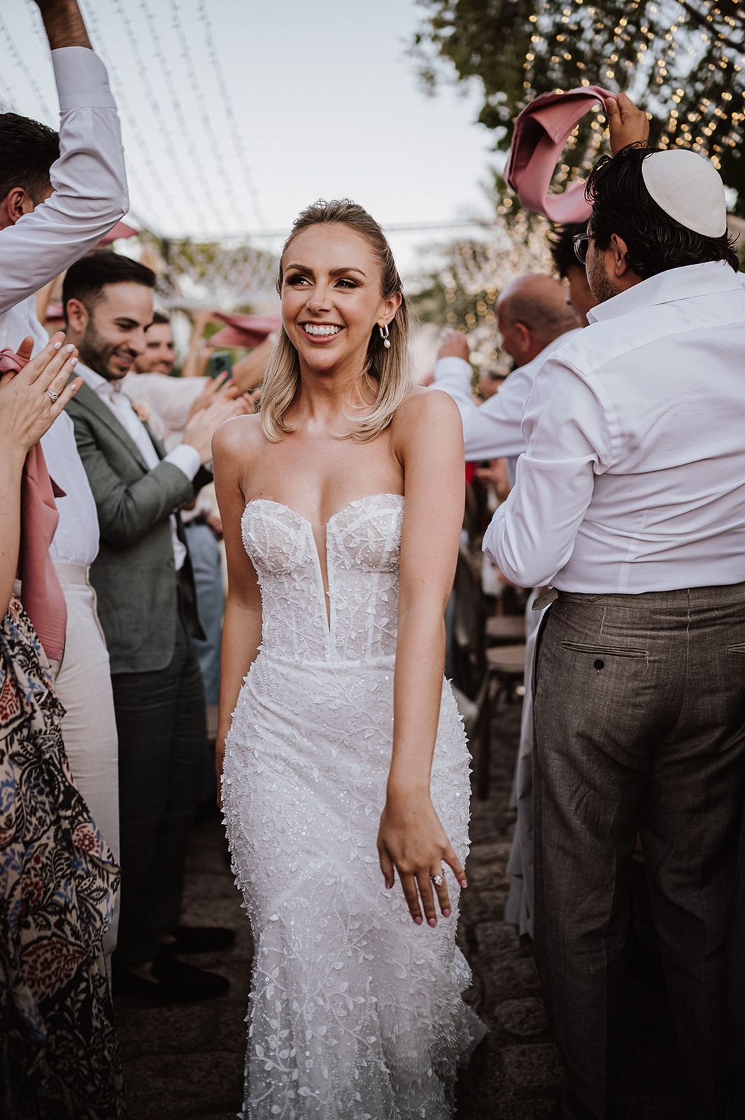 Radiant bride in strapless lace gown smiles joyfully surrounded by wedding guests.