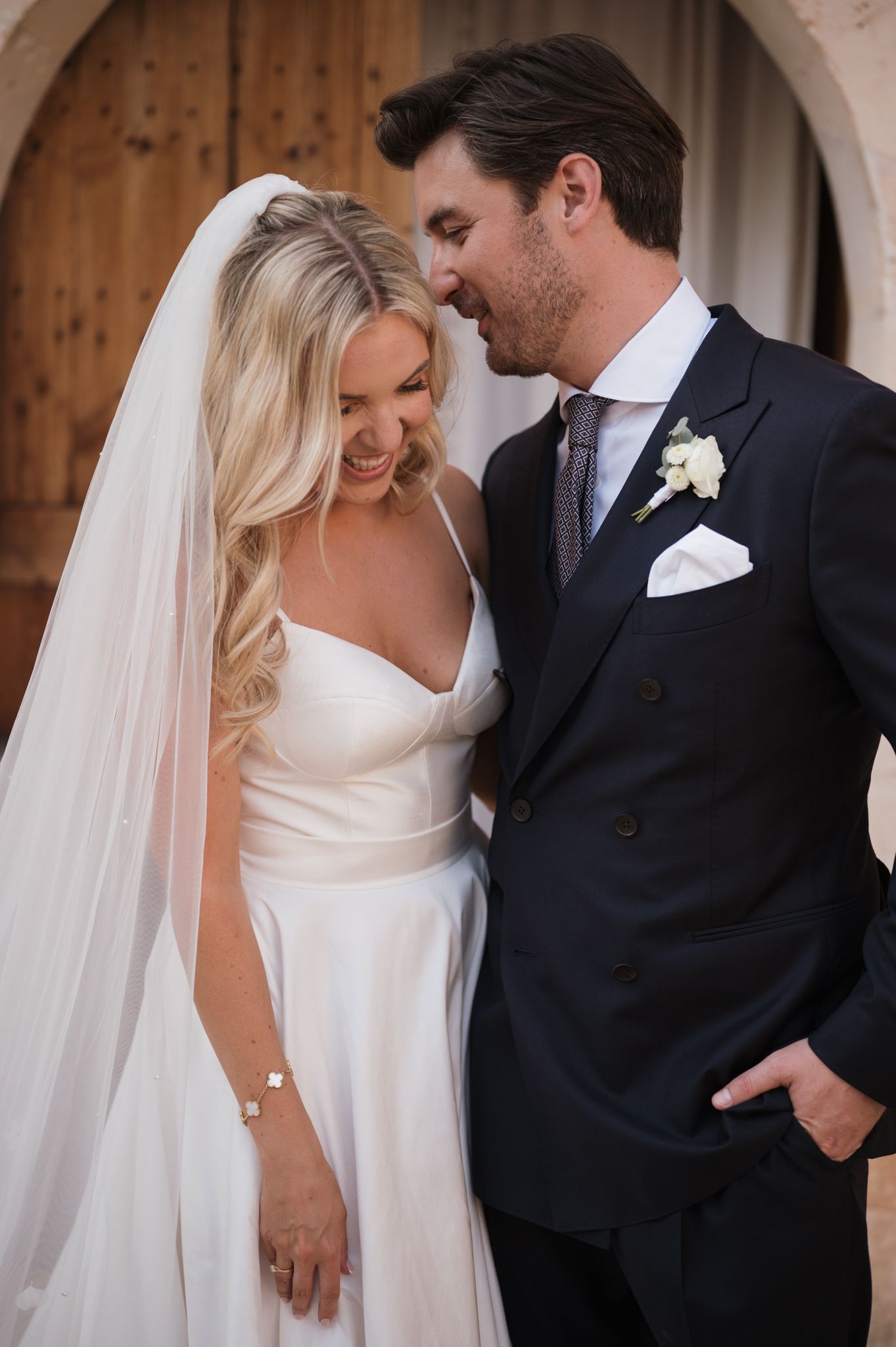Bride in white satin gown and veil smiles with groom in navy suit during romantic outdoor wedding portrait.