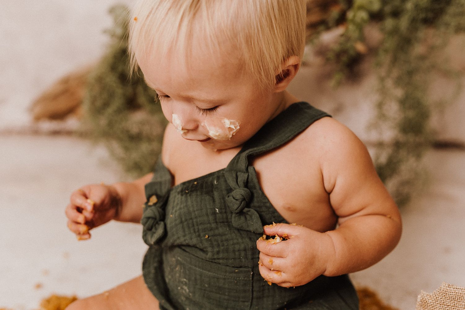 A messy toddler in a green romper enjoys cake smash outdoors, with frosting on their face.